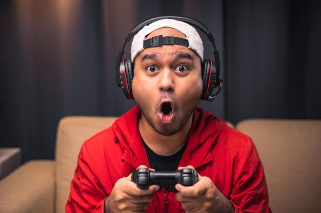 Excited and shocked face of Asian gamer with headphone holding joystick playing video game online sitting on sofa at living room Indian professional gamer streaming on social playing game very fun