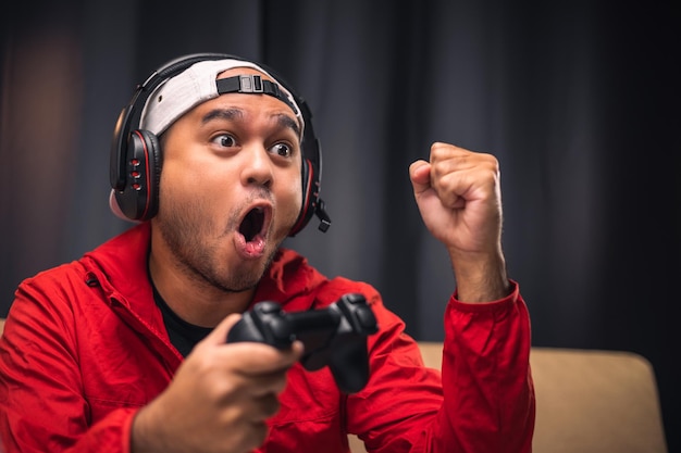 Excited and shocked face of Asian gamer with headphone holding joystick playing video game online sitting on sofa at living room Indian professional gamer streaming on social playing game very fun