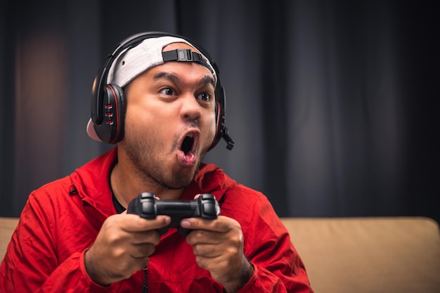 Excited and shocked face of Asian gamer with headphone holding joystick playing video game online sitting on sofa at living room Indian professional gamer streaming on social playing game very fun