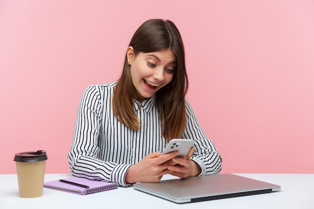 Взволнованная шокированная женщина-офисный работник, удивленная распродажами и скидками, делающая покупки в Интернете на своем смартфоне, сидя на рабочем месте. Съемка в помещении студии на розовом фоне