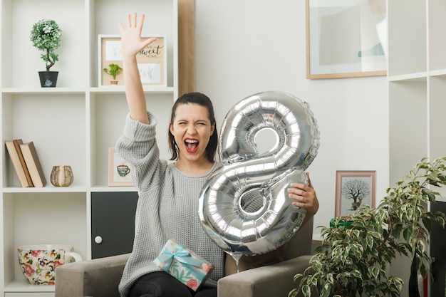 Excited showing five beautiful girl on happy women day holding number eight balloon sitting on armchair in living room