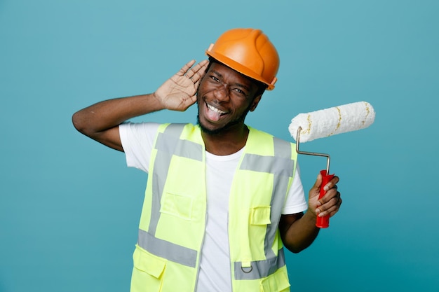 Mostra eccitata ascolta il gesto giovane costruttore afroamericano in uniforme che tiene una spazzola a rullo isolata su sfondo blu