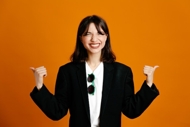 Excited showing thumbs up young beautiful female wearing black jacket isolated on orange background