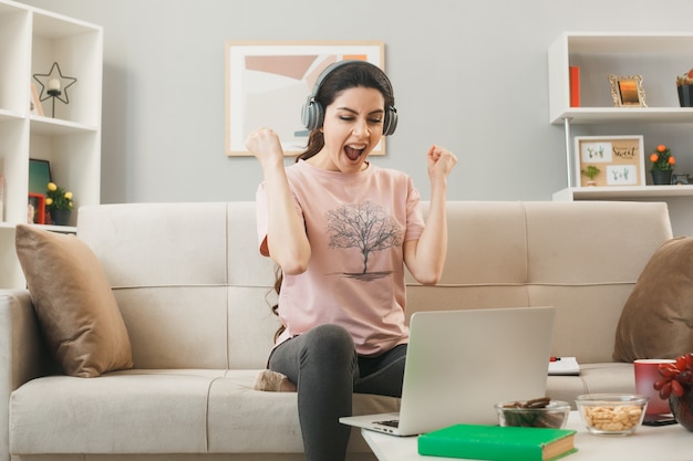 Excited showing yes gesture young girl wearing headphones used laptop sitting on sofa behind coffee table in living room