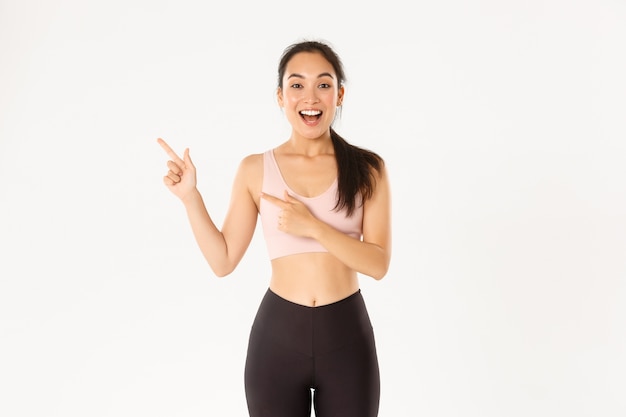   Excited slim and cute asian fitness girl, female athlete pointing fingers upper left corner, showing workout equipment sale promo or gym offer.