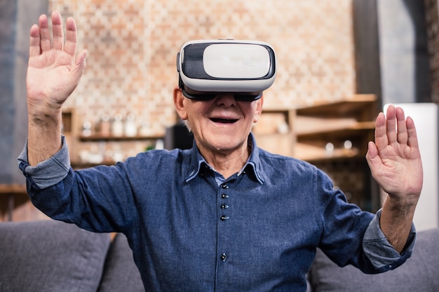 Excited smilin elderly man sitting at home in VR glasses and expressing joy