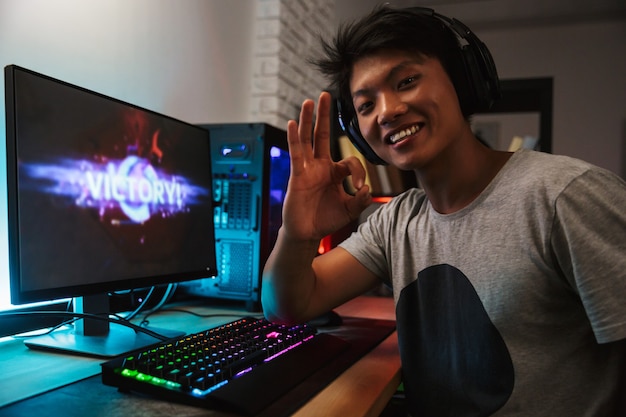 Excited smiling gamer boy rejoicing victory while playing video games on computer in dark room, wearing headphones and using backlit colorful keyboard