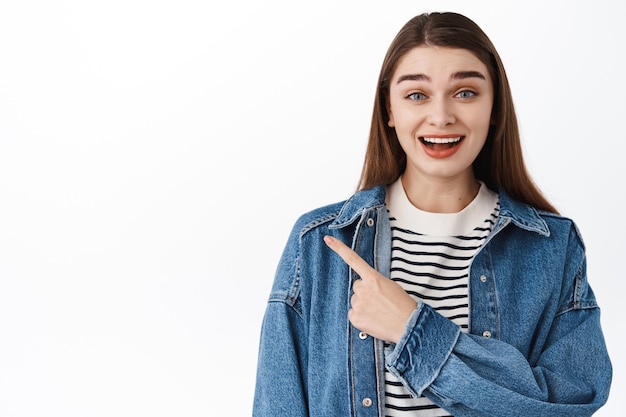 Взволнованная улыбающаяся девушка-студентка, указывающая в сторону слева с рекламным текстом, показывая пальцем баннер с логотипом, с интересом смотрит, стоит над белой стеной