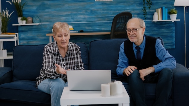 Excited smiling senior couple looking at laptop computer waving during videocall sitting at couch. Cheerful retired husband and wife laughing talking at virtual meeting resting on sofa in cozy room.