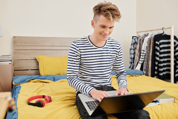 Excited smiling teenage boy sitting on bed, working on laptop and making post for his blog when spending time in bedroom during quarantine