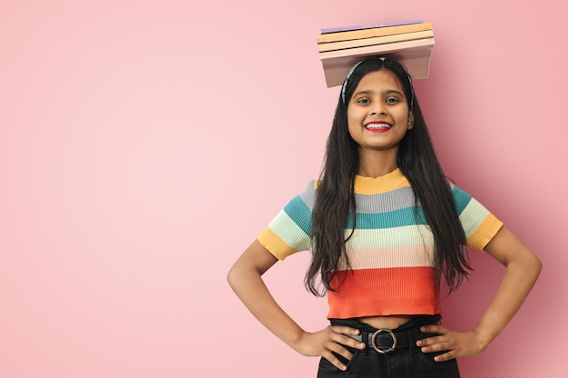 Excited smiling young indian asian girl student standing straight with her hands on waist posing islolated balancing books on her head