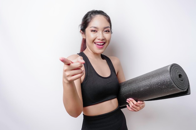 Excited sporty Asian woman wearing sportswear pointing at the camera while carrying yoga mat isolated by white background