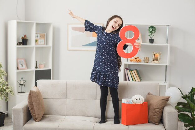 Excited spreading hand little girl on happy women's day holding number eight standing on sofa in living room