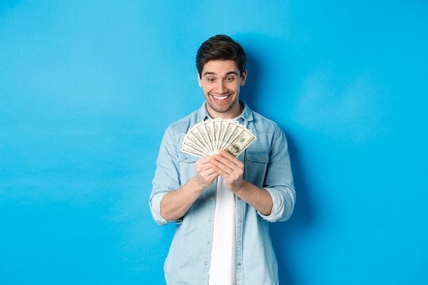 흥분된 성공적인 남자는 돈을 세고, 현금에 만족하고 웃고, 파란색 배경 위에 서 있습니다.