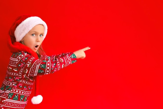 Excited surprise kid girl wearing Christmas red Santa hat green pajamas costume isolated red studio background with finger pointed up sale and discount Shopping Banner copy space for text mock up