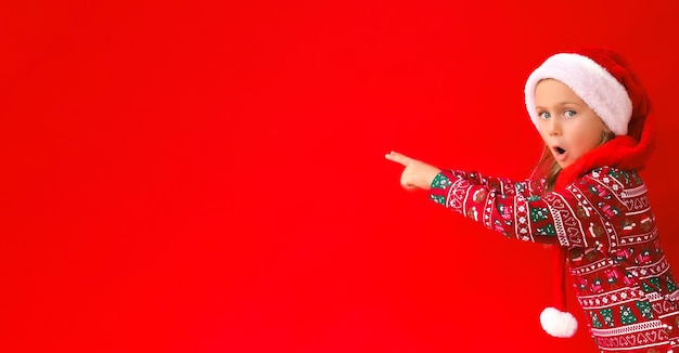 クリスマスの赤いサンタ帽子緑のパジャマ コスチュームを身に着けている興奮した驚きの子供の女の子は指で指を指して赤いスタジオ背景を分離しました。
