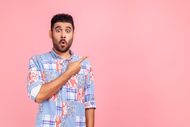 Excited surprised handsome man with beard wearing blue casual shirt looking at camera with open mouth, pointing finger aside, showing copy space. Indoor studio shot isolated on pink background.