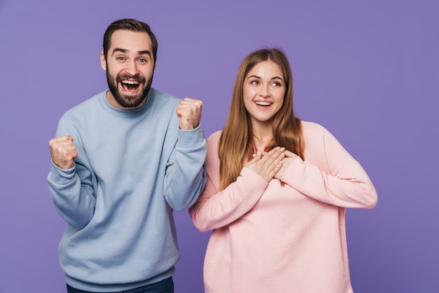 excited surprised positive loving couple isolated over purple wall make winner gesture.