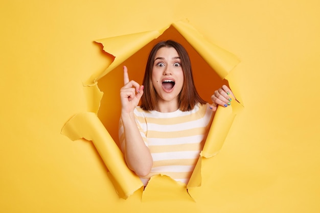 Excited surprised smart young adult woman stands in torn paper hole having great idea looking at camera with raised finger looking through breakthrough of yellow background