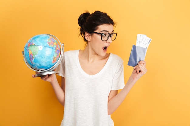 Excited surprised woman wearing glasses holding globe and passport with tickets.