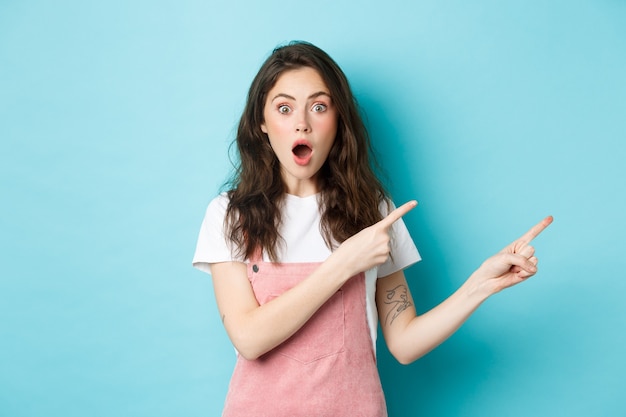 Excited and surprised young woman open mouth, say wow and pointing fingers right at logo, staring impressed at camera while talking about new product or news, blue background