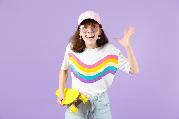 Excited teen girl in vivid clothes, eyeglasses hold skateboard,\
keeping mouth open spreading hands isolated on violet pastel\
background. people sincere emotions, lifestyle concept. mock up\
copy space.