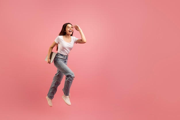 Excited teen girl with laptop computer in hand jumping on pink background