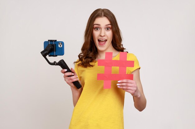 Excited teenage girl with phone on stabilizer leads videoblog, looking at camera with overjoyed expression, showing red hashtag symbol. Indoor studio shot isolated on gray background.