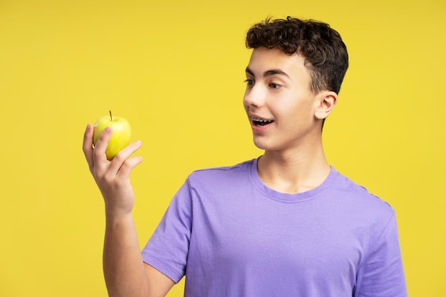 Excited teenager boy holding apple fruit looking at him standing isolated on yellow background