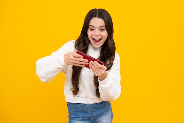 Excited teenager girl use smart phone share social media\
chatting by mobile phone wear stylish casual trendy clothes\
isolated over yellow background child with smartphone