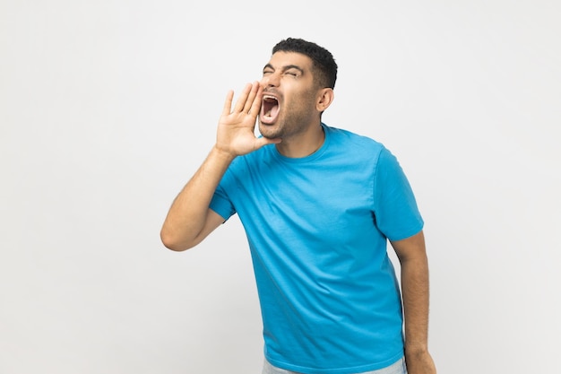 Возбужденный небритый мужчина в синей футболке стоит и держит руки возле открытого рта, крича новости