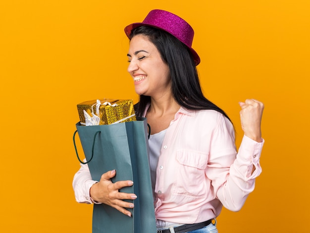 Eccitato con gli occhi chiusi, giovane bella ragazza che indossa un cappello da festa che tiene in mano una borsa regalo che mostra un gesto di sì