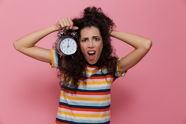 excited woman 20s with curly hair holding alarm clock isolated on pink