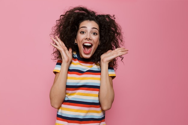 excited woman 20s with curly hair shouting and throwing up hands isolated on pink