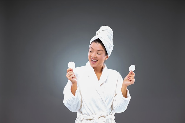 Excited woman in a bathrobe and a towel wrapped around her head holds a cotton white pad while standing in front of a gray wall