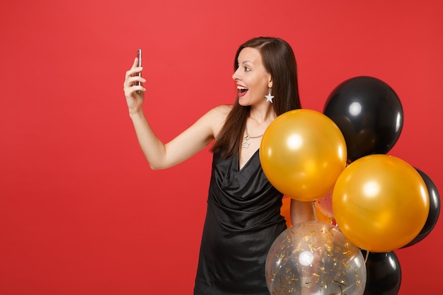 Excited woman in black dress holding air balloons doing taking selfie shot, making video call, talking on mobile phone isolated on red background. Happy New Year birthday mockup holiday party concept.