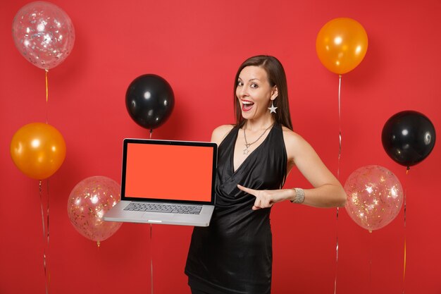 Excited woman in black dress pointing index finger on laptop pc computer with blank black empty screen on bright red background air balloons. Happy New Year, birthday mockup holiday party concept.