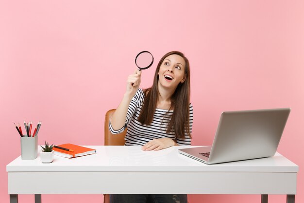 Excited woman in casual clothes looking up through magnifying glass work at white desk with contemporary pc laptop isolated on pastel pink background. Achievement business career concept. Copy space.