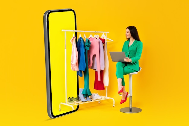 Excited woman fashion designer with laptop looking at clothes on rail near huge cellphone on yellow