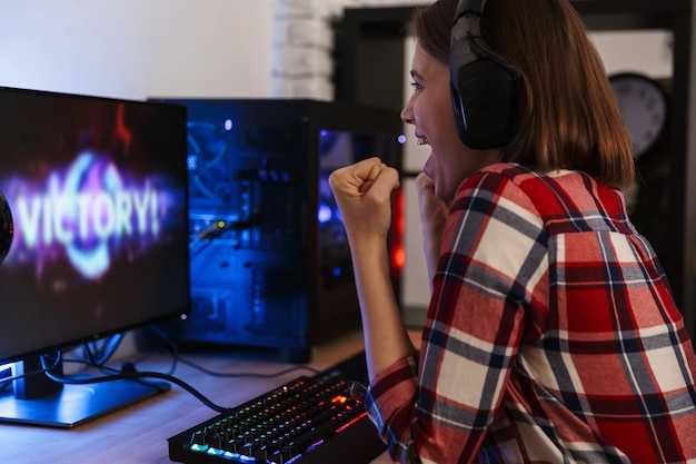 Excited woman gamer sitting at the table, playing online games on a computer indoors, celebrating victory