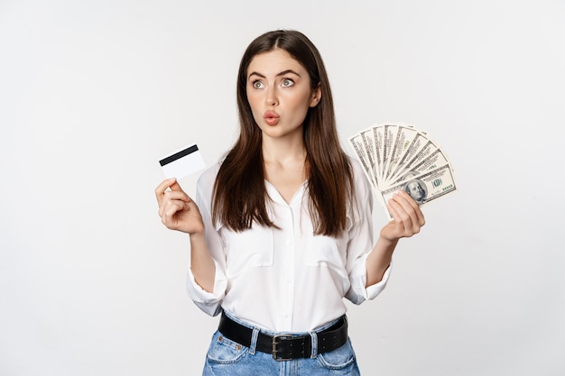 Excited woman holding credit card and money, looking amazed at cash, standing against white background