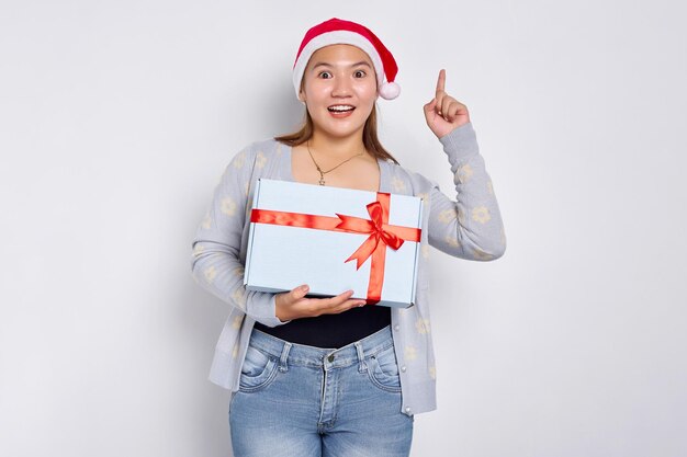 Excited woman holding in hand a Christmas gift Beautiful Asian woman in Santa Claus hat pointing finger up isolated on white studio background Merry Christmas Concept