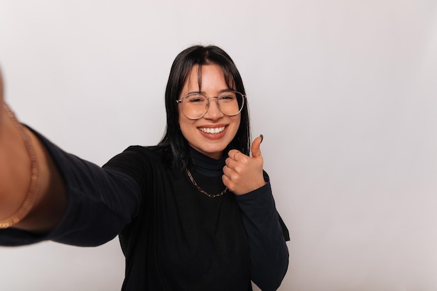 Excited woman is taking a selfie while showing thumb up over white backdrop