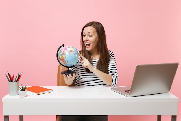 Excited woman looking on world globe planning vacation while sit and work at white desk with contemporary pc laptop isolated on pastel pink background. Achievement business career concept. Copy space.