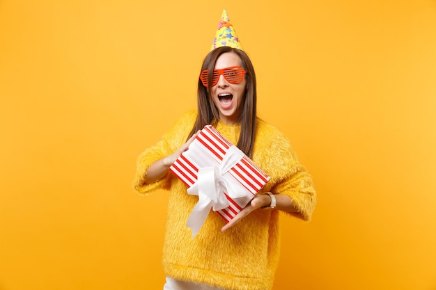 Excited woman in orange funny glasses, birthday hat holding red box with gift present celebrating and enjoying holiday isolated on bright yellow background. People sincere emotions, lifestyle concept.