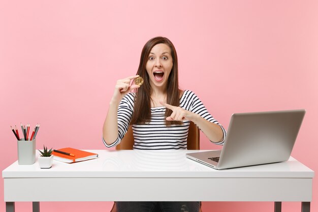 Excited woman pointing index finger on bitcoin, metal coin of golden color, future currency sit work at white desk with pc laptop isolated on pink background. achievement business career. copy space