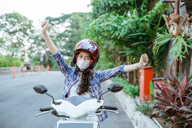 Excited woman raises both hands using helmet and mask on motorbike with road background