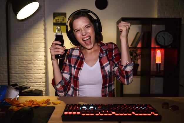 Excited woman wearing headset playing online games on computer, eating snacks