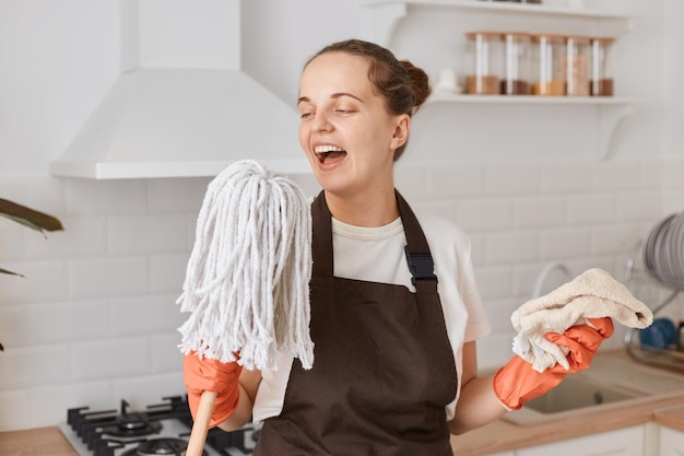 Foto donna eccitata con i capelli scuri che indossa una maglietta bianca e un grembiule marrone che tiene un mocio e un panno in mano governante che canta mentre lava la sua cucina divertendosi mentre pulisce la casa