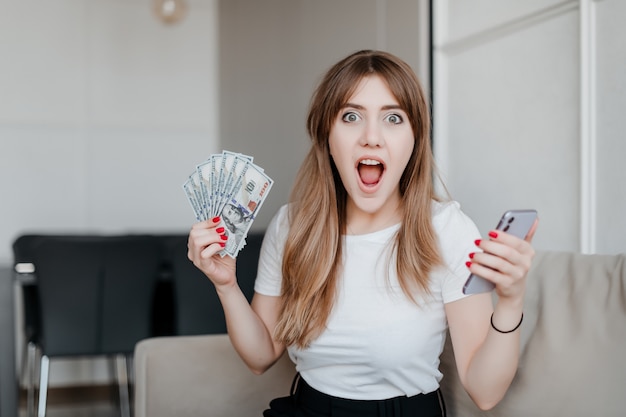 Excited woman with dollar bills money and phone in hand sitting on couch at home